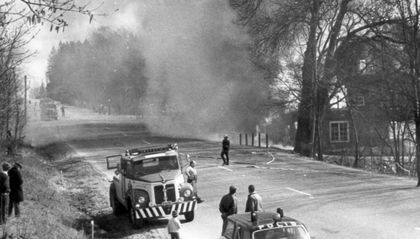 Svartvit äldre bild med brandbil och polis vid vägen och en rykande fastighet.