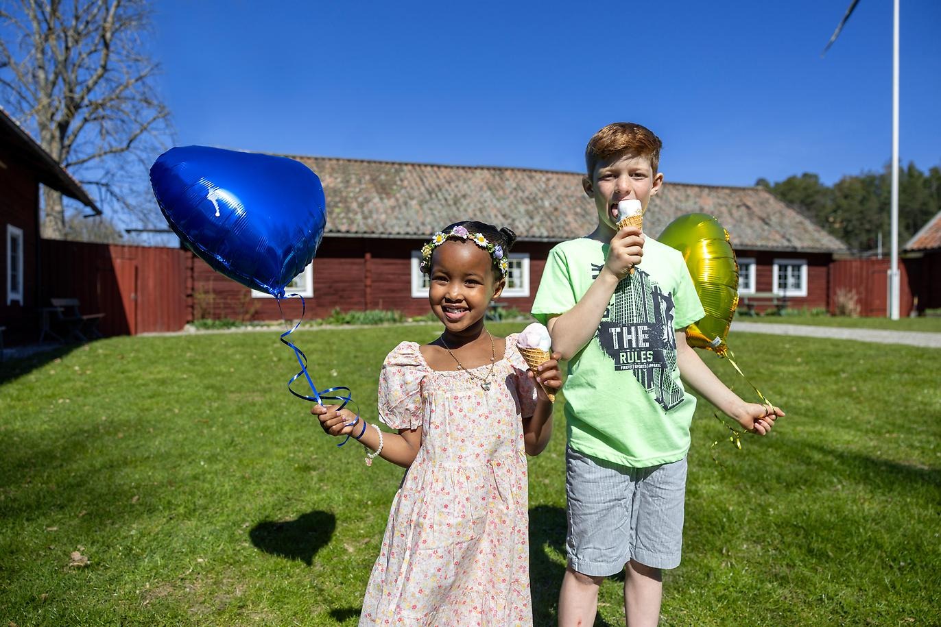 En flicka och en pojke med varsin gul ch blå ballong i handen. Framför en äldre gårdsbyggnad.