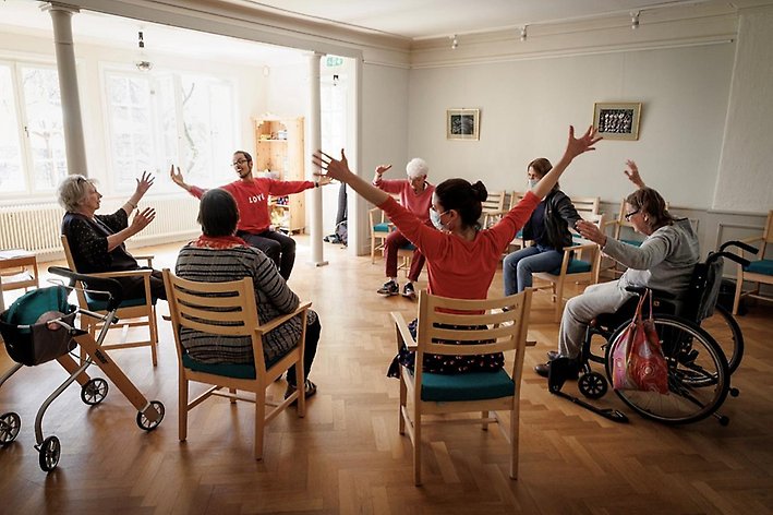 Man leder rörelseaktivitet för äldre och personal sittandes i stolar och rullstolar på äldreboende