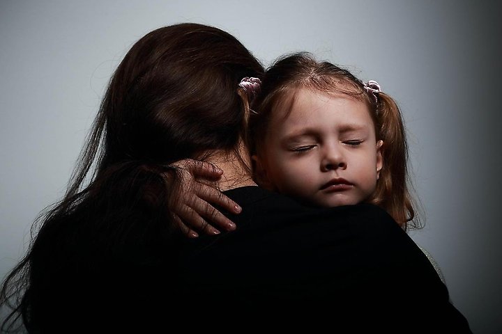 Litet barn som blundar och kramar sin mamma, man ser ryggen på mamman och ansiktet på barnet