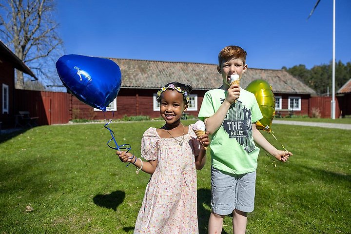 En flicka och en pojke med varsin gul ch blå ballong i handen. Framför en äldre gårdsbyggnad.